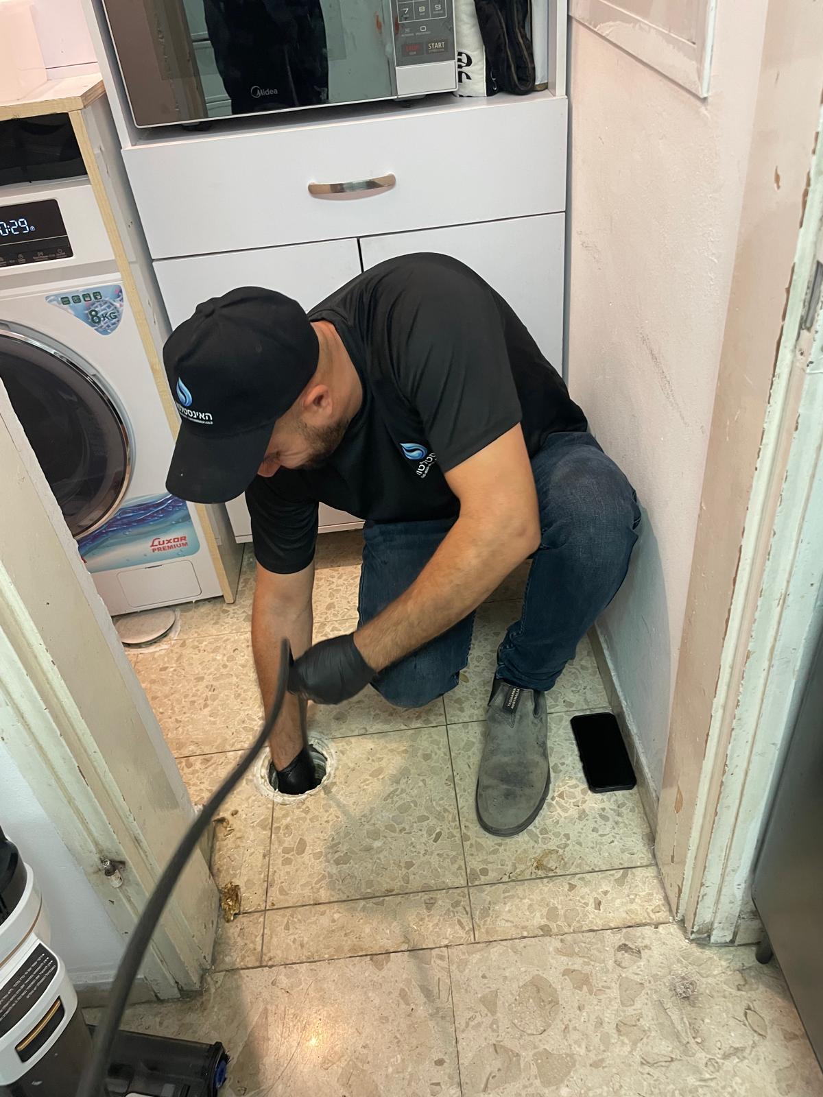 יצחק האינסטלטור מבצע שטיפת קו ביוב בלחץ גבוה דרך פתח ניקוז ברצפה ליד מכונת כביסה - חולון, מאי 2026